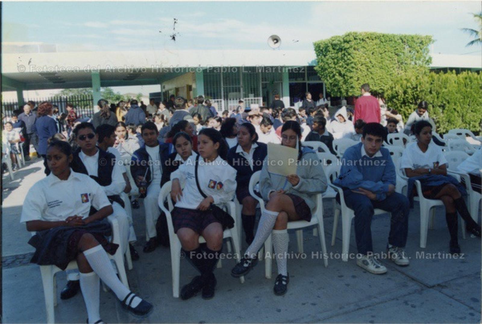 Alumnos sentados en sillas acomodadas en explanada de escuela.