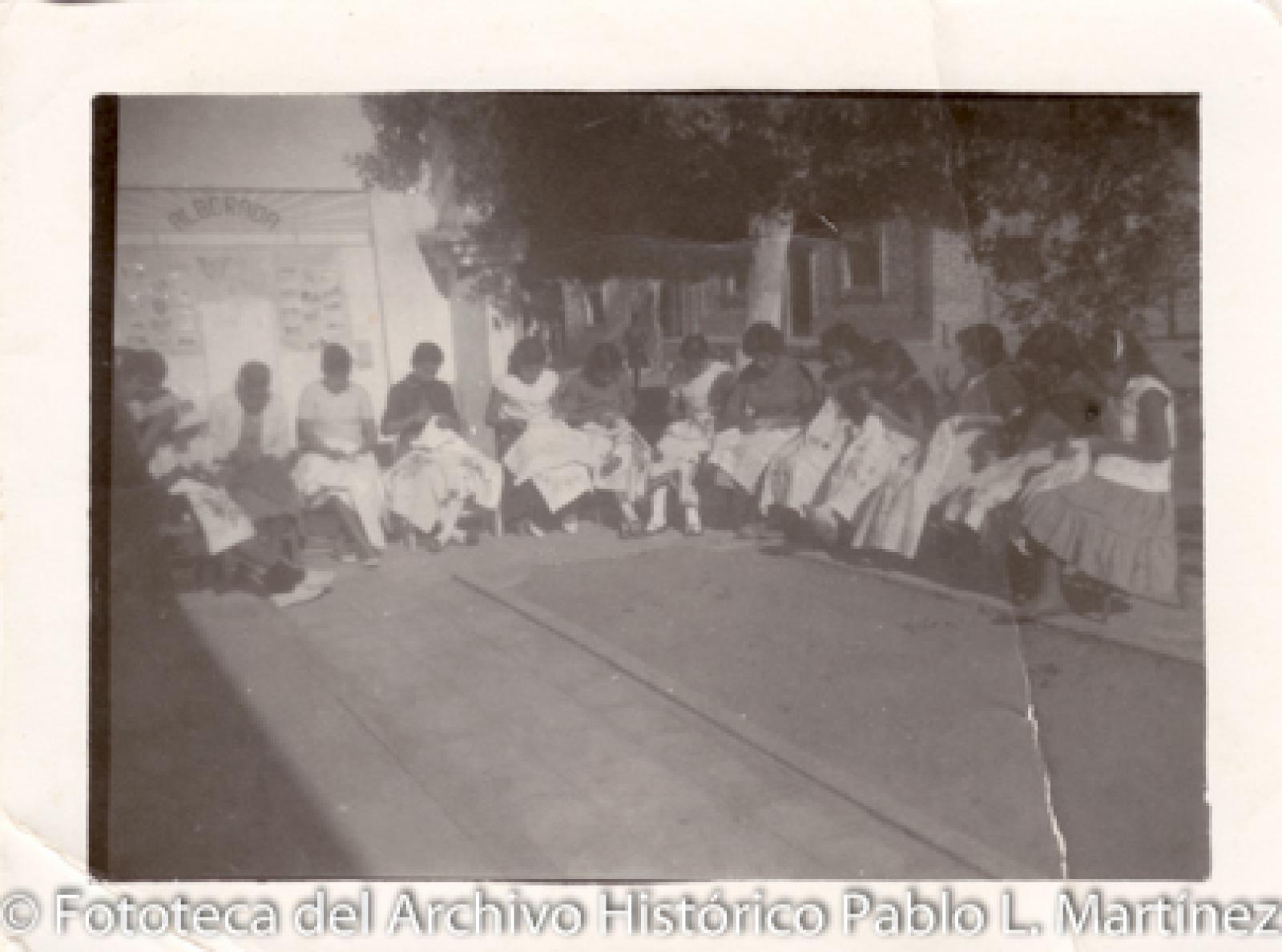 Grupo de mujeres que bordan al aire libre frente a árboles y casa de ladrillo.