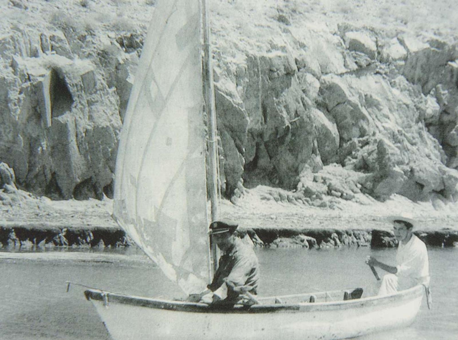 Hombres que navegan en velero, frente a playa rocosa.