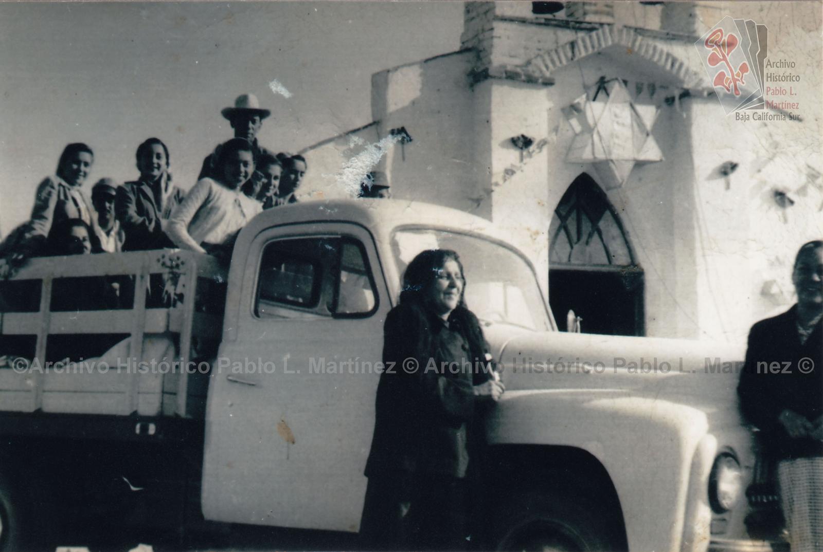 Hombres y mujeres en camión, a su espalda el templo a María Auxiliadora, en Comondú, Baja California.