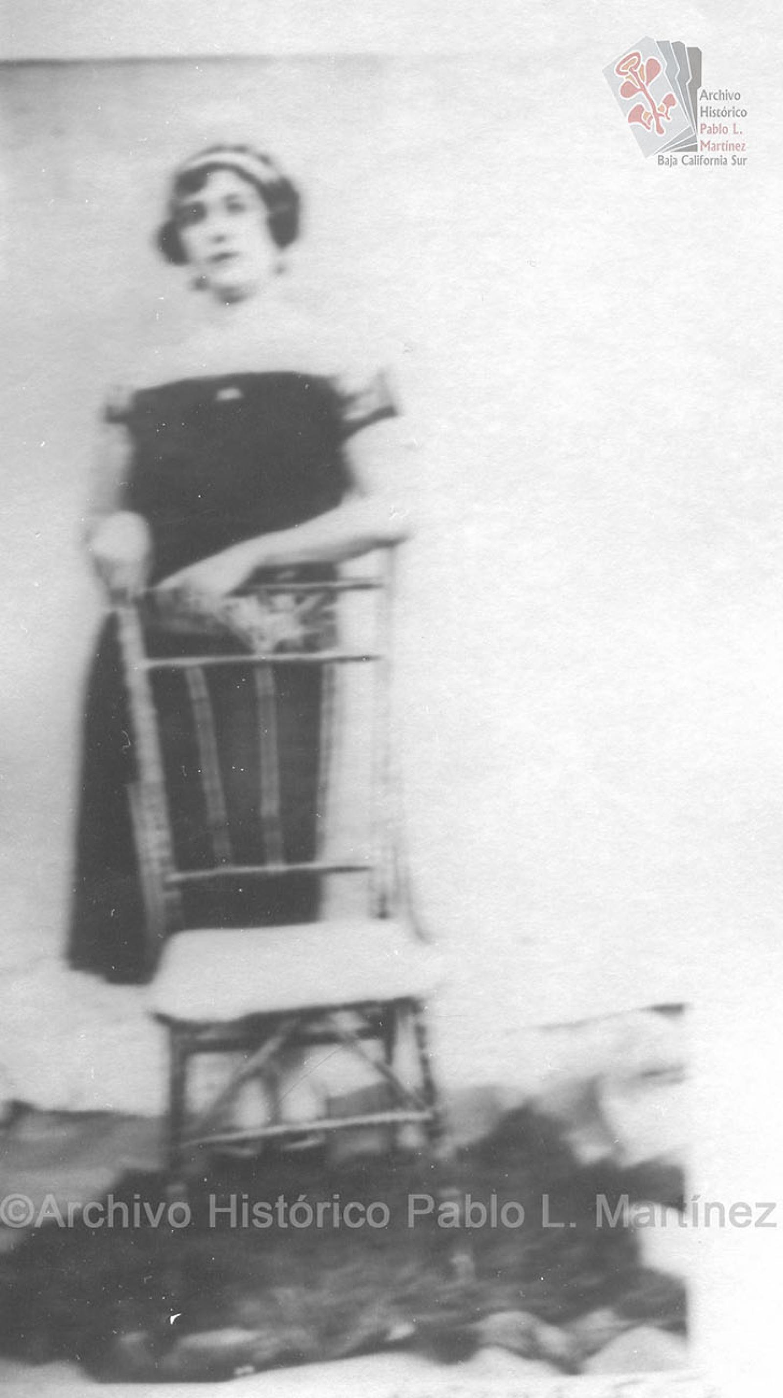 Mujer de pie se apoya en silla, viste elegante.