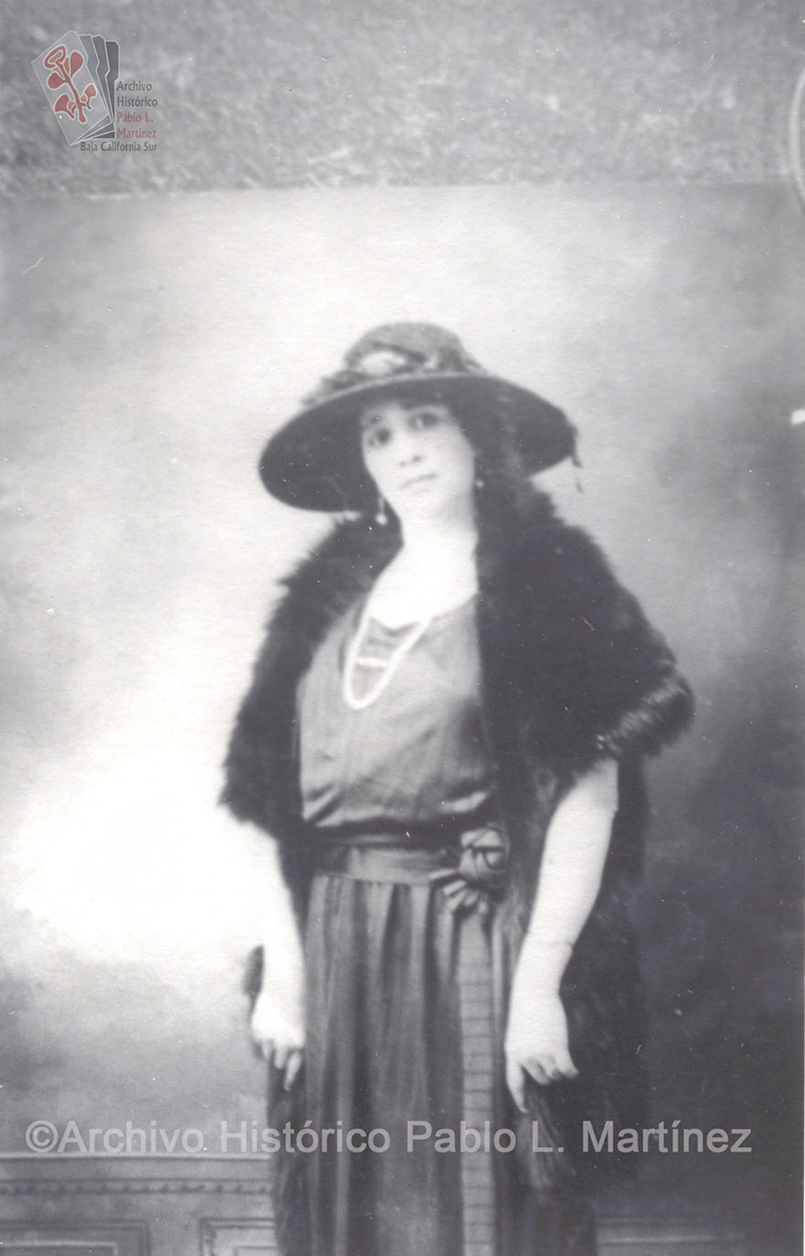 Mujer que posa de pie, adorna con un sombrero su vestuario elegante.