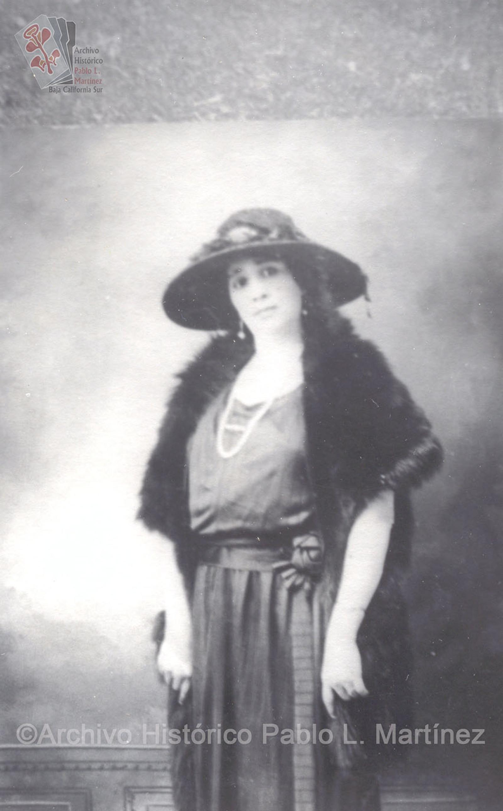 Mujer que posa de pie, viste elegante y porta sombrero.