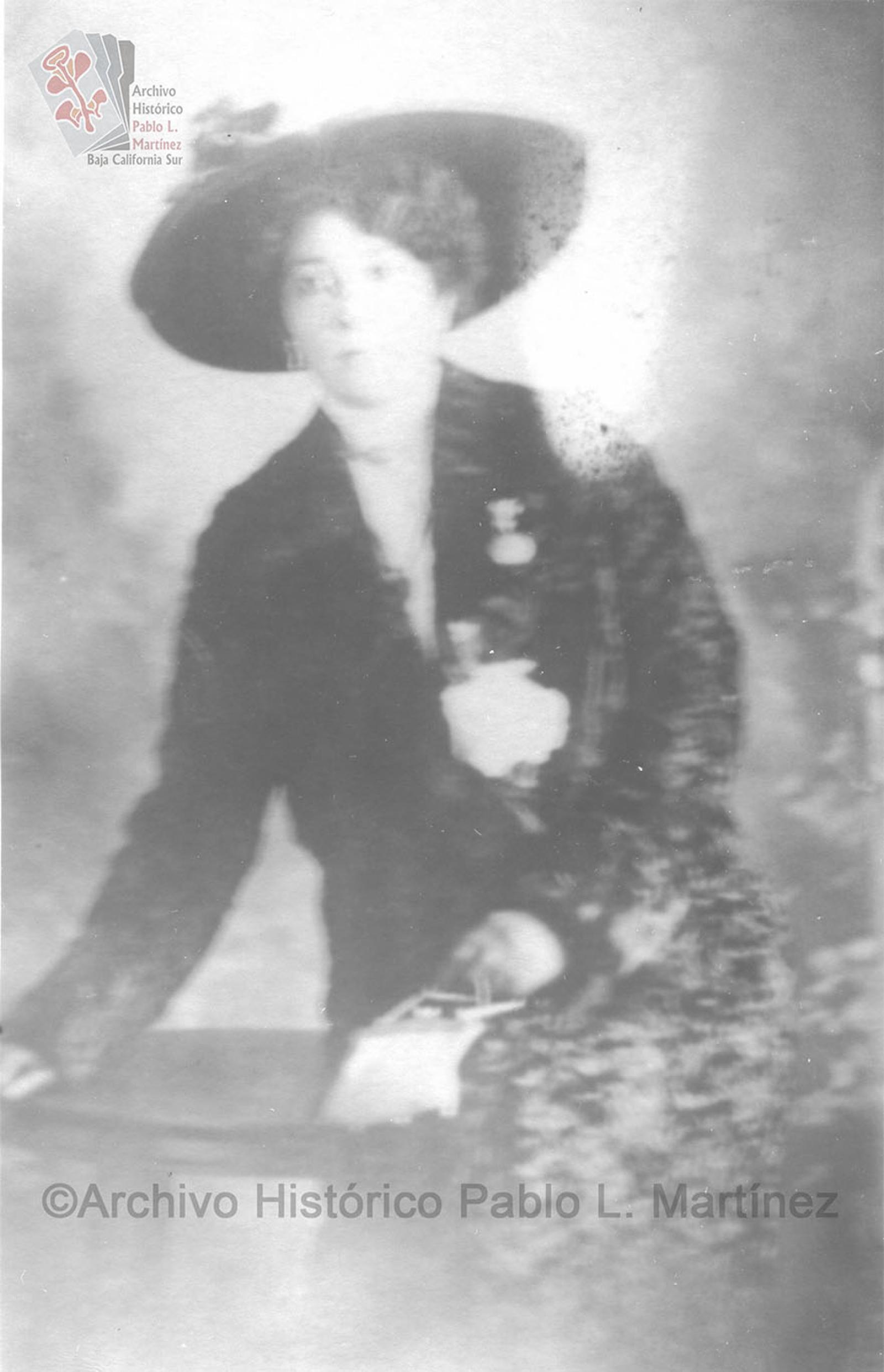 Mujer que posa en una mesa, viste elegante y porta sombrero.
