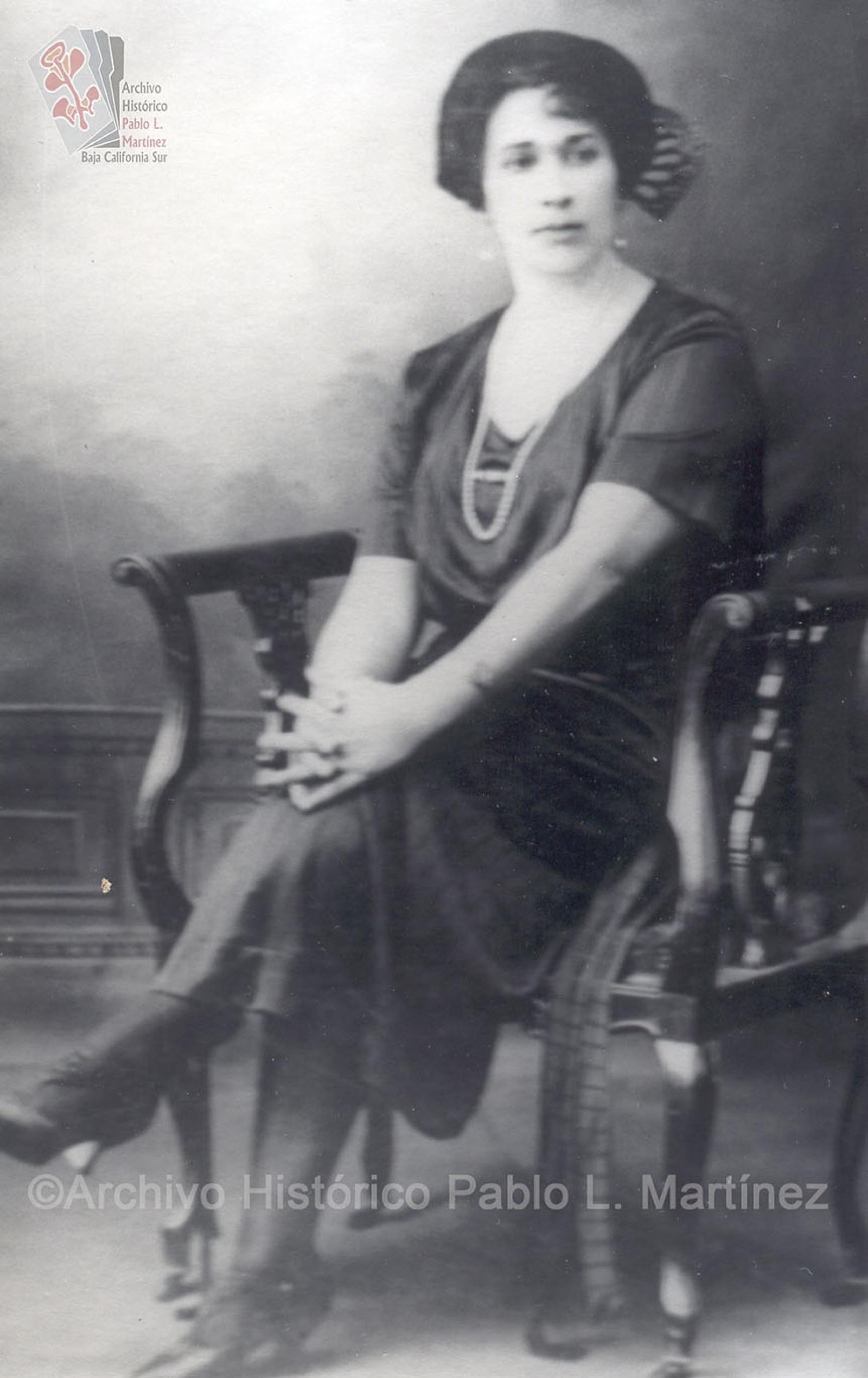 Mujer que posa sentada con sus manos entrelazadas, viste atuendo elegante.