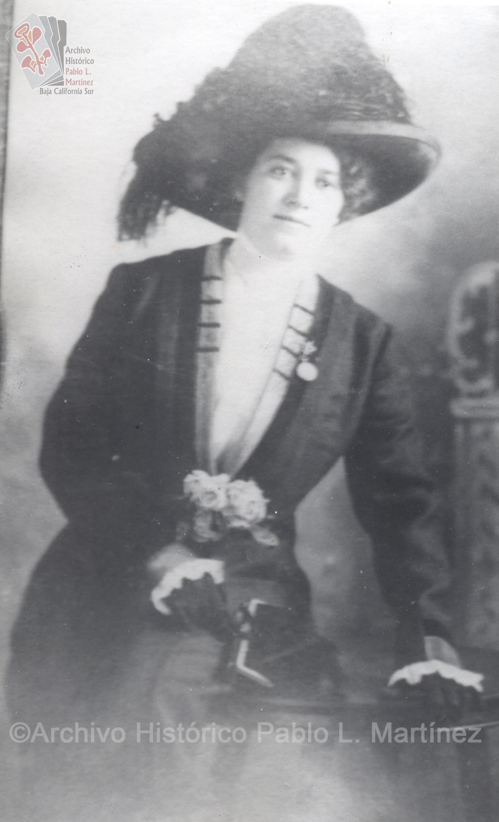 Mujer que posa sentada en una mesa, viste elegante y porta sombrero con plumas.