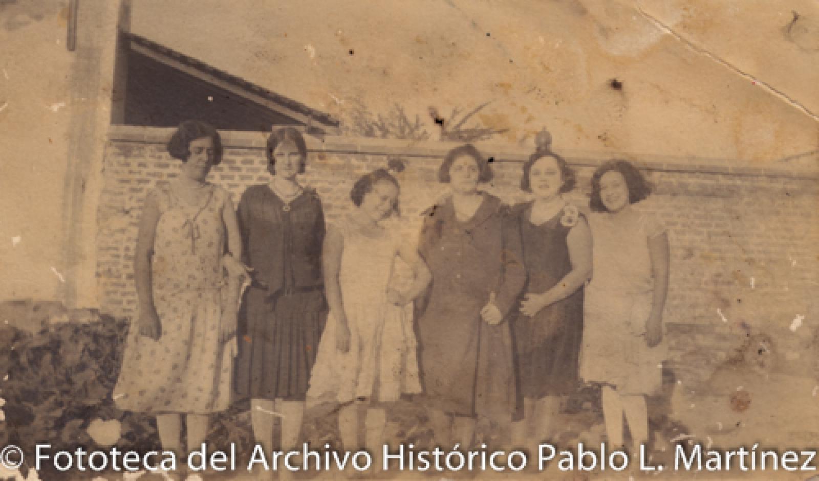 Mujeres, frente a barda de ladrillo.