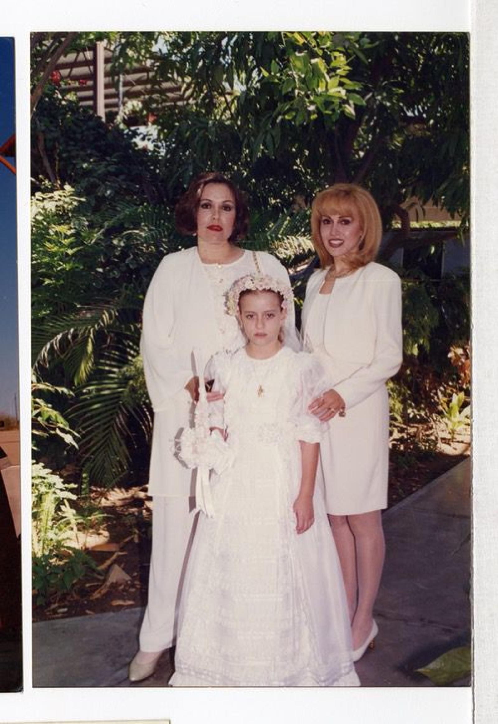 Niña con vestuario de Primera Comunión acompañada de dos mujeres, retrato.