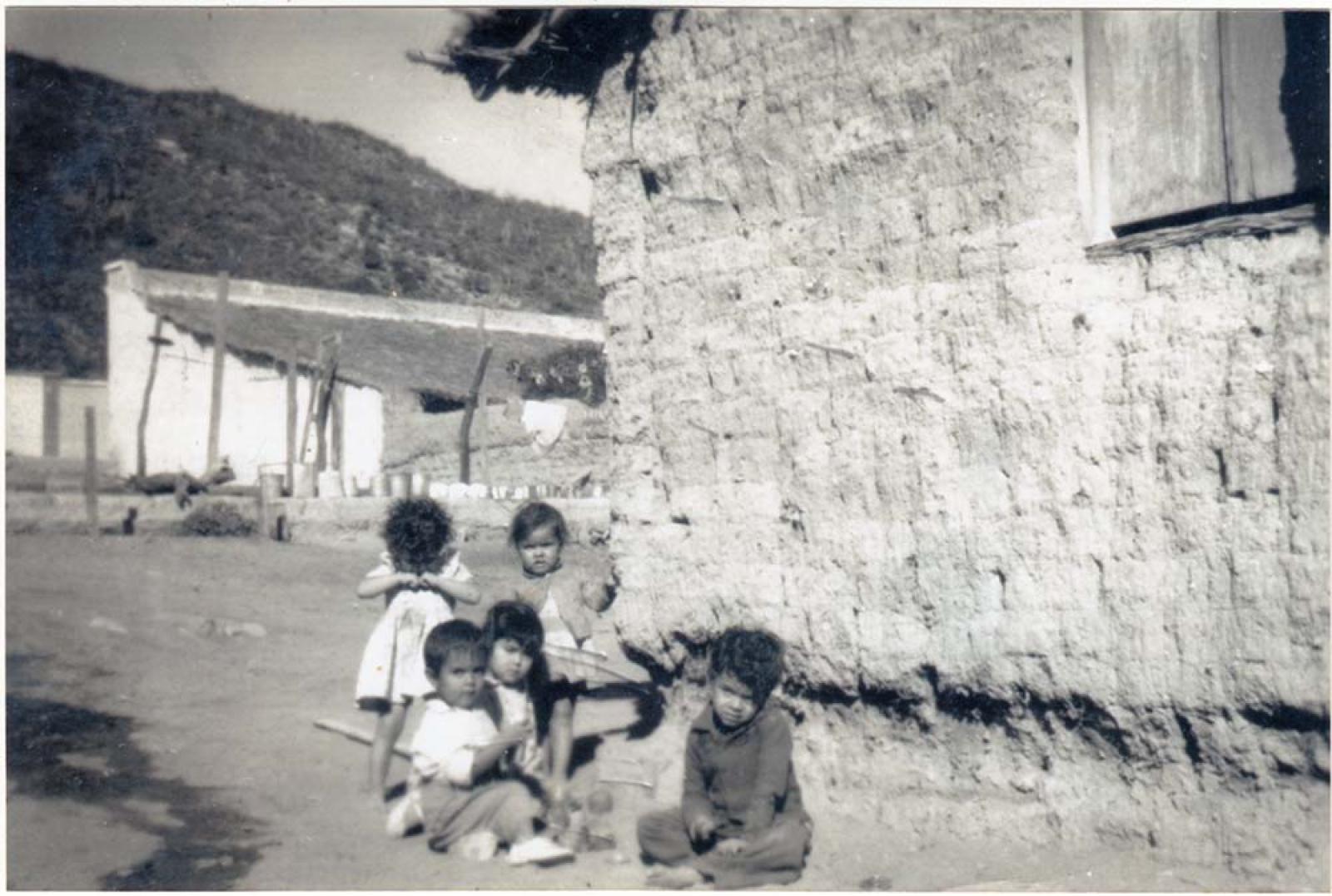 Niños y niñas juegan junto a casa de adobe y palma.