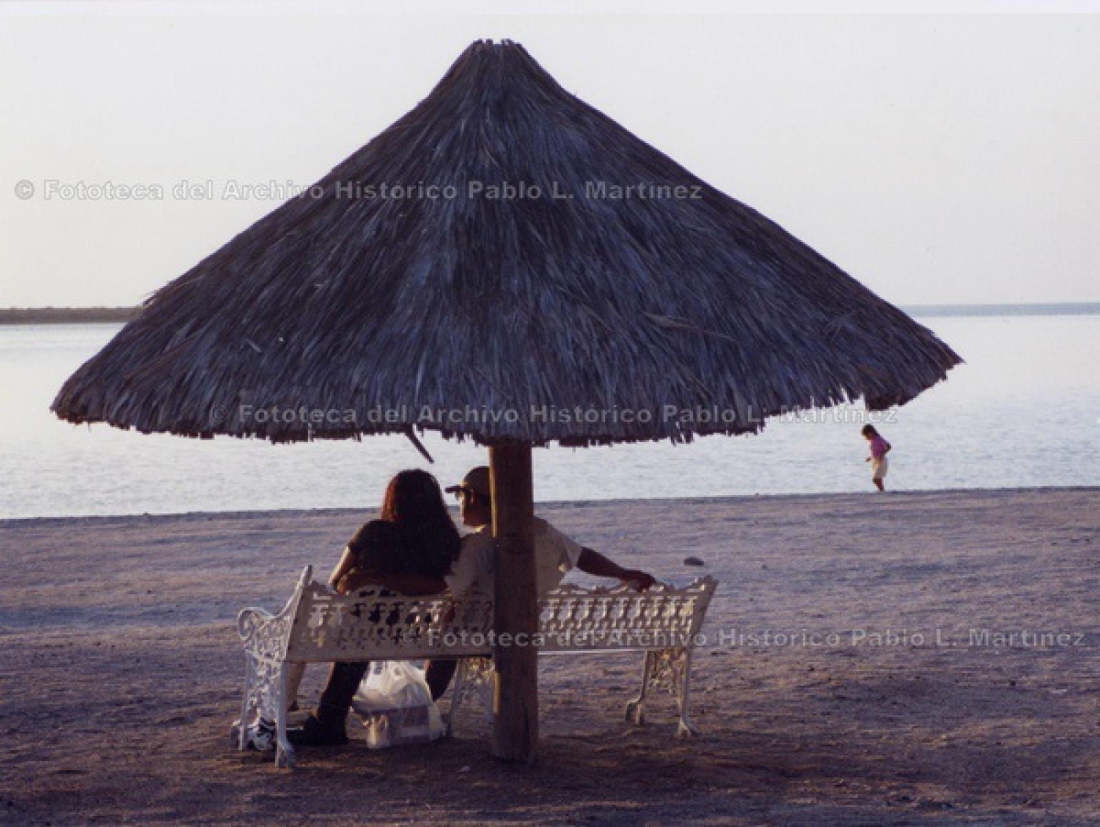 Pareja admira el mar, sentados en banca bajo palapa.